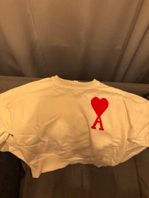 Vit tröja med rött hjärta från Ami - Vit tröja från Ami med ett rött hjärta och bokstaven 'A' broderat på bröstet. Tröjan har rund halsringning, ribbade muddar och är gjord i mjukt material. Enkel och stilren design som sticker ut med den ikoniska loggan.
