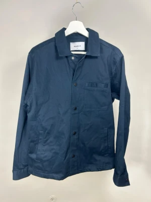 Woodbird Overshirt | S - Stilren mörkblå overshirt från Woodbird med klassisk krage och knappstängning framtill. Jackan har en bröstficka och två sidofickor. Tillverkad i ett mjukt bomullstyg som passar perfekt för lager på lager. Enkel och clean design som funkar till det mesta.