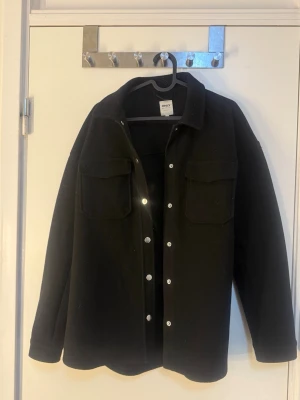 Svart overshirt från ONLY - Stilren svart overshirt från ONLY med klassisk krage, tryckknappar framtill och två stora bröstfickor. Overshirten har en rak passform och är tillverkad i ett tjockare tyg som ger en snygg och avslappnad look. 