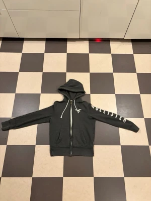 Mörkgrå zip hoodie från Hollister - Mörkgrå hoodie från Hollister med dragkedja framtill, vita snören i huvan och tryckt logga på ena ärmen. Två fickor fram och ribbade muddar. Perfekt för en avslappnad och cool stil. Vintage/acrhive hollister