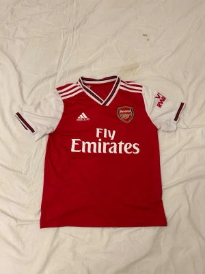 Arsenal matchtröja Adidas röd/vit - Snygg Arsenal matchtröja från Adidas i rött och vitt med klubbmärke på bröstet och Fly Emirates-tryck. Tröjan har vita ärmar med röda detaljer och klassiska Adidas-ränder på axlarna. Perfekt för dig som älskar fotboll och vill stötta Arsenal.