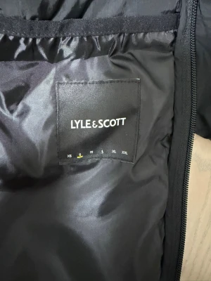 Svart pufferjacka från Lyle & Scott - Säljer en svart pufferjacka från Lyle & Scott. Jackan är tillverkad i 100% polyamid med vaddering och foder i polyester. Klassisk design med dragkedja och diskret logga på insidan. Perfekt för kalla vinterdagar. Använd några gånger 