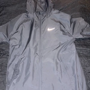 Grått Nike träningsset med huva - Säljer ett grått träningsset från Nike med jacka och byxor. Jackan har huva, dragkedja och två fickor framtill samt en vit Nike-logga på bröstet. Byxorna har resår i midjan och en matchande vit logga på benet. Perfekt för träning eller chill.