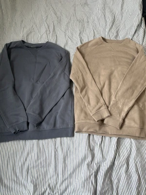Två basic crewneck sweatshirts - Säljer två stilrena crewneck sweatshirts i dämpade färger, en i gråblått och en i beige. Båda har klassisk passform, ribbade muddar och rund halsringning. Perfekta för en avslappnad och enkel look. Materialet känns mjukt och bekvämt, troligen bomull. Båda för 100