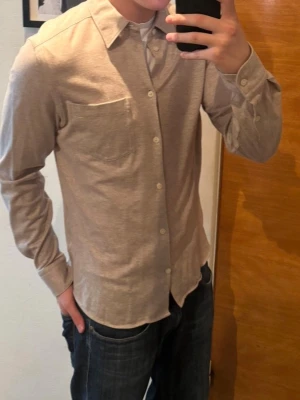 Beige J.LINDBERG skjorta i bomull med knappar - Stilren beige skjorta i mjuk bomull med klassisk krage och vita knappar framtill. Skjortan har en bröstficka och lång ärm, perfekt för en clean och enkel look. Passformen är normal och materialet känns bekvämt mot huden.