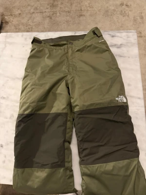 Olivgröna täckbyxor från The North Face - Täckbyxor i olivgrönt från The North Face med förstärkta mörkgröna partier på knäna och en unik bakficka med camomönster och orange detaljer. Byxorna har rak passform och är perfekta för kalla dagar utomhus.                                                              Storlek L i barn