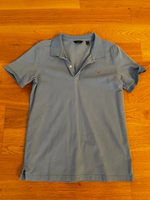  Piké - Snygg ljusblå polotopp från GANT med klassisk krage och två knappar framtill. T-shirten har ett broderat GANT-emblem på bröstet och är gjord i mjuk bomull. Perfekt för dig som gillar stilrena och bekväma plagg.