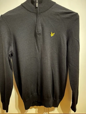 Svart halvzip-tröja från Lyle & Scott - Stilren svart stickad tröja från Lyle & Scott med halv dragkedja och hög krage. Tröjan har det klassiska gula logobroderiet på bröstet och ribbade muddar vid ärmslut och nederkant. Perfekt för lager på lager och enkel att matcha.