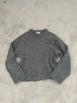 Grå stickad tröja  - En enkel och stilren grå stickad tröja från gina  med rund halsringning och ribbade muddar vid ärmslut och nederkant. Tröjan har en rak passform och är tillverkad i mjukt material som passar perfekt för kyliga dagar.