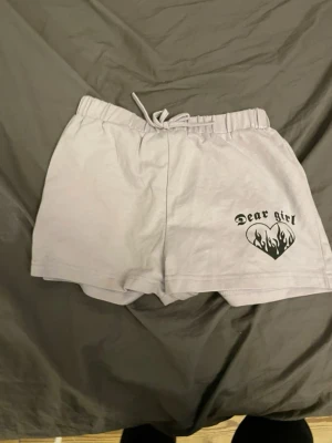Ljusgrå shorts med tryck och snörning - Säljer ett par lila shorts med snörning i midjan och ett svart tryck med texten 'Dear girl' och ett hjärta med flames på ena benet. Sköna och casual, perfekta för sommaren.