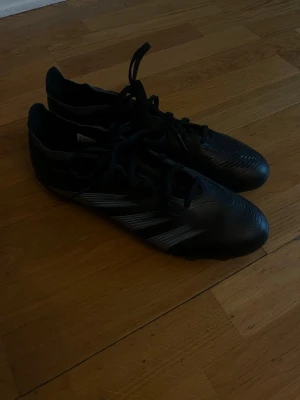 Svarta Adidas fotbollsskor - Snygga svarta fotbollsskor från Adidas med klassiska vita ränder på sidan. Skorna har snörning och dobbar för bra grepp på planen. Tillverkade i syntetmaterial som ger en lätt och smidig känsla när du spelar. Använda 1-2 gånger 