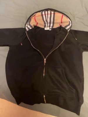 Svart hoodie med dragkedja och mönstrad huva - Svart hoodie med dragkedja från Burberry. Klassisk huva fodrad med det ikoniska rutmönstret i beige, rött och svart. Mjukt material och ribbade muddar. Perfekt för en stilren och avslappnad look.