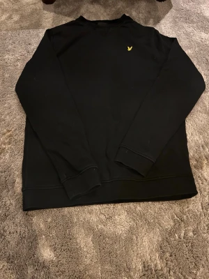 Svart sweatshirt från Lyle & Scott - Svart sweatshirt med rund hals från Lyle & Scott. Tröjan har ett broderat gult örnlogga på bröstet och långa ärmar. Perfekt för en stilren och avslappnad look.
