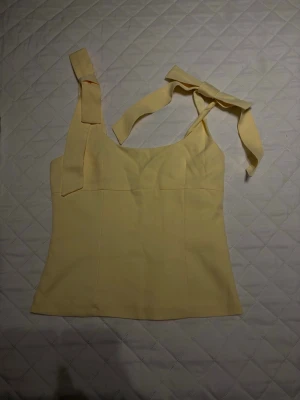 Gul linnetopp med knytband - Söt ljusgul linnetopp med breda axelband som knyts till rosetter. Toppen har en enkel, figurnära passform och ärmlös design. Perfekt för varma dagar och ger en fräsch look till din outfit.