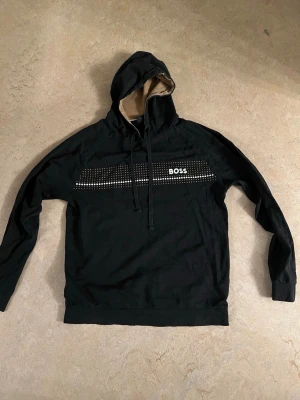 Svart hoodie från BOSS - Svart hoodie från BOSS med vit logga och mönstrad detalj över bröstet. Huva med beige insida och dragsnören. Mjuk bomullskänsla och klassisk passform, perfekt för en avslappnad stil.
