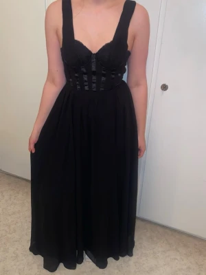 Svart långklänning - Elegant svart långklänning med breda axelband och markerad byst. Livet har dekorativa banddetaljer och kjolen faller mjukt i ett svepande tyg. Perfekt för en speciell kväll eller bal. Klänningen är ärmlös och har en unik look.