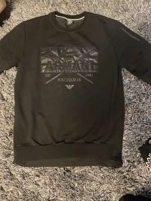 Svart sweatshirt från Armani Exchange - Snygg svart sweatshirt från Armani Exchange med glansigt tryck och broderad logga på bröstet. Tröjan har ribbade muddar vid ärmslut och nederkant samt coola detaljer på ärmarna. Perfekt för dig som gillar stilrena och exklusiva plagg.