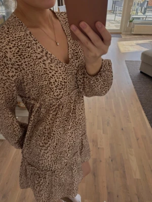 Beige kortklänning med v-ringning - Säljer en beige kortklänning med brunt prickigt mönster och v-ringad hals. Klänningen har långa ärmar och volangdetalj nedtill. Perfekt för dig som gillar en avslappnad men trendig stil. Perfekt till stranden också! 