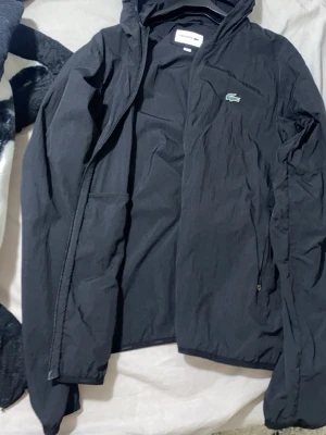 Svart vindjacka från Lacoste - Snygg svart vindjacka från Lacoste med dragkedja framtill och huva. Jackan har två fickor med dragkedja och den klassiska gröna krokodilloggan på bröstet. Tillverkad i lätt och tåligt material, perfekt för blåsiga dagar.