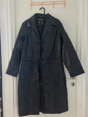 Svart lång kappa i croc-mönster Gatto - Snygg svart lång kappa från Gatto med croc-mönstrad yta och klassisk krage. Kappan har knäppning framtill och två stora fickor. Materialet har en glansig finish som ger en cool look. Perfekt för dig som vill sticka ut i höst och vinter.