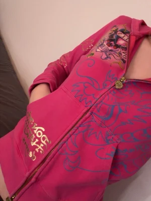 Ed Hardy zipper - Rosa ziphoodie från Ed Hardy i strl M. Jackan har gulddetaljer på armarna och luvan. Jackan har tryck på ryggen med stenar och tryck på framsidan med två fickor. 