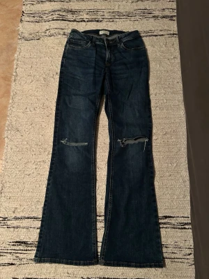 Mörkblå bootcut jeans med slitningar - Säljer ett par mörkblå bootcut jeans med slitna detaljer över knäna. Jeansen har klassisk femficksmodell, låg midja och vida ben. Perfekta för dig som gillar en avslappnad och trendig look.