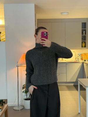 Mörkgrå stickad tröja med halvzip - Mysig mörkgrå stickad tröja med ribbad struktur och halvzip i halsen. Tröjan har hög krage och långa ärmar, perfekt för att hålla värmen. Materialet ser ut att vara ull eller en ullblandning, vilket gör den extra varm och skön.