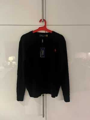 Svart tröja från Polo Ralph Lauren - Klassisk svart tröja från Polo Ralph Lauren med rund halsringning och den ikoniska röda logotypen broderad på bröstet. Långärmad modell i mjukt stickat material som passar perfekt till jeans eller kjol. Tröjan är storlek M men lite mindre i storlek. 