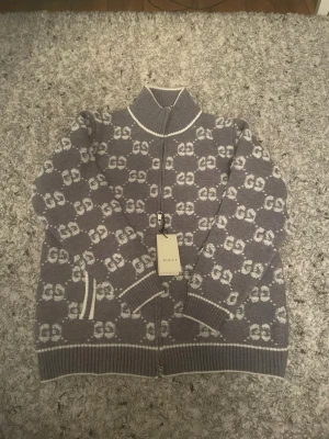 Grå ziptröja med GG-mönster från Gucci - Snygg grå ziptröja från Gucci med vitt GG-monogram över hela plagget. Tröjan har ribbade muddar, hög krage och två fickor framtill med vita detaljer. Tillverkad i mjukt stickat material och har en lyxig känsla. Perfekt för dig som gillar exklusiva streetwear-vibbar.