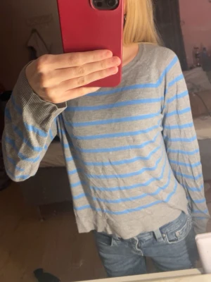 Grå tröja med blå ränder - Säljer en grå långärmad tröja med tunna blå horisontella ränder. Tröjan har rund halsringning och ribbade muddar vid ärmslut och nederkant. Perfekt för dig som gillar en enkel och avslappnad stil.