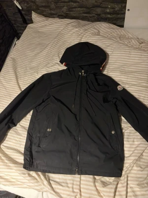 Svart Moncler windbreaker - Snygg svart vindjacka från Moncler med justerbar huva och dragkedja framtill. Jackan har två sidofickor med tryckknappar och Moncler-logga på ärmen. Tillverkad i lätt, vattenavvisande material som passar perfekt för blåsiga dagar. En metall del av snöret har ramlat av och den är lite smutsig!