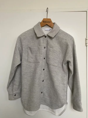 Ljusgrå overshirt från Name It - Stilren ljusgrå overshirt från Name It köpt på Åhlens, med två bröstfickor och svarta knappar framtill. Skjortan har klassisk krage och är tillverkad i ull. Perfekt lager på lager-plagg för en avslappnad look.