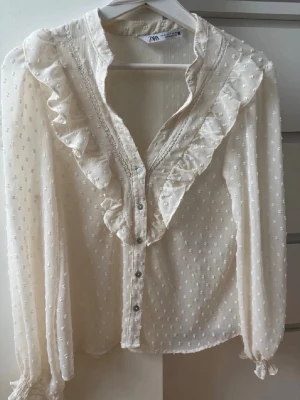 Vit volangblus från Zara - Söt vit blus do Zara med volangdetaljer längs ringningen och knappar framtill. Blusen har diskret prickigt mönster i tyget och transparent känsla. Långa ärmar med resår vid handlederna ger en romantisk vibe. Perfekt till jeans eller kjol.