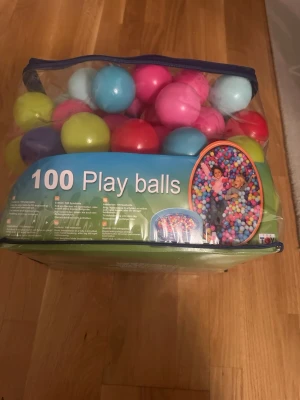 100 färgglada lekbollar i väska - Säljer en genomskinlig väska fylld med 100 lekbollar i olika färger som blå, rosa, röd, lila och gul. Perfekt för bollhav eller lek i barnrummet. Bollarna är gjorda av plast och kommer i en praktisk förvaringsväska med dragkedja.