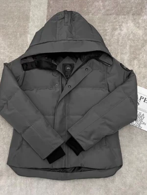 Mörkgrå pufferjacka med huva - Snygg mörkgrå pufferjacka från Canada Goose med stor huva och diskret logotyp på ärmen. Jackan har dragkedja och tryckknappar framtill samt ribbade muddar vid ärmsluten. Perfekt för kalla vinterdagar och riktigt varm tack vare det vadderade materialet.