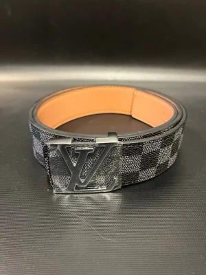 Rutmönstrat Louis Vuitton bälte - Snyggt bälte från Louis Vuitton med klassiskt rutmönster i svart och grått. Bältet har en stor LV-logga i silver på spännet och insidan är i ljust skinn. Perfekt accessoar för att lyfta din outfit.