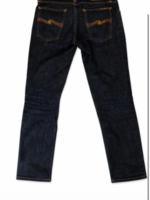 Mörkblå jeans från Nudie Jeans - Säljer ett par mörkblå jeans från Nudie Jeans med klassisk rak passform. Jeansen har kontrastsömmar och ikoniska broderade bakfickor. Tillverkade i kraftig denim med fem fickor och knappgylf. Perfekta för dig som gillar stilrena och tidlösa jeans.