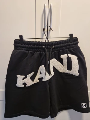 Svarta shorts från Karl Kani - Svarta shorts från Karl Kani med stor vit logga i frotté på framsidan. Shortsen har resårmidja med snörning och en liten logotyp nertill. Perfekta för en avslappnad och streetinspirerad stil.