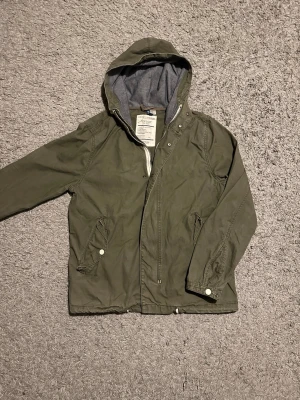 Olivgrön vindjacka med huva - Snygg militärgrön vårjacka med grått innerfoder i huvan. Jackan har dragkedja och tryckknappar framtill, två snedställda fickor med lock och tryckknapp samt justerbara ärmslut. Perfekt för blåsiga dagar. Den är i riktigt fint skick, 8/10. Skriv vid minsta lilla intresse 🙌