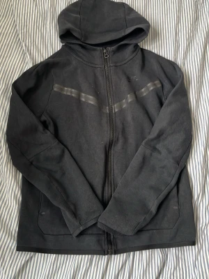 Svart Nike tech - Snygg svart hoodie med dragkedja framtill och huva. Hoodien har två diskreta fickor och ett grått randigt mönster över bröstet. Tillverkad i mjukt bomullsmaterial, perfekt för en avslappnad stil.