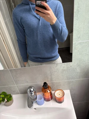 Blå hoodie i mjukt material - Säljer en stilren blå hoodie med huva och dragsnören. Hoodien har en smal passform och är gjord i ett mjukt, stickat material som känns skönt mot huden. Perfekt för dig som gillar en enkel men snygg look. Långa ärmar och ribbade muddar vid ärmslut och nederkant. Vill ni ha mer bilder bara hör av er.