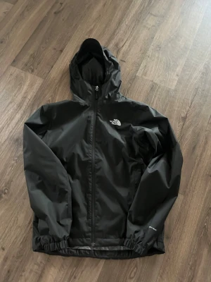Svart regnjacka från The North Face - Snygg svart regnjacka från The North Face med huva och dragkedja framtill. Jackan har elastiska muddar vid ärmsluten och logga på bröstet och ryggen. Tillverkad i vattenavvisande material, perfekt för regniga dagar.