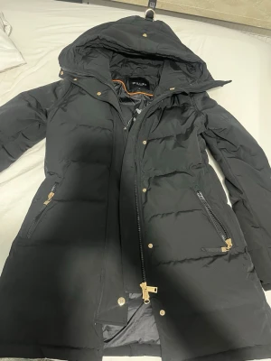 Svart lång dunjacka från Hollies - Snygg svart dunjacka från Hollies med huva och guldfärgade dragkedjor och knappar. Jackan är lång, har två framfickor med dragkedja och quiltad design. Perfekt för kalla dagar och riktigt varm. Klassisk och stilren look. Denna jackan köpte jag i december 2025. Jag tog av pälsen då det går att ta av och sätta på när man vill. Pälset är såklart äkta, och pälset är svart. Jag beställde den från Hollies hemsida och den kostade 6.249kr. Jag har använt den ett par gånger men den är fortfarande väldigt