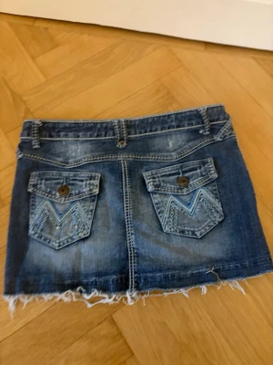 Blå jeanskjol med fransig kant - Säljer en blå jeanskjol med fransig nederkant och slitna detaljer. Kjolen har fickor både fram och bak, bälteshällor och snygga dekorsömmar på bakfickorna. Perfekt för dig som gillar en avslappnad och trendig stil.