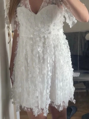 Vit kortklänning med spets och blommor - Söt vit kortklänning med 3D-blommor och spetsdetaljer. Klänningen har korta ärmar. Perfekt för sommarens alla tillfällen och studentfirande.