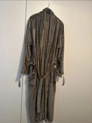 Mjuk brun fleece morgonrock - Säljer en supermjuk och varm brun morgonrock i fleece. Den har långa ärmar, två stora fickor framtill och ett matchande knytband i midjan. Perfekt för att mysa runt hemma under kyliga dagar.
