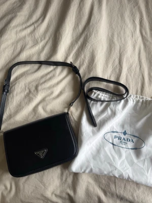 Svart axelväska - Stilren svart axelväska i slätt läder med justerbar axelrem och logga på framsidan. Väskan har en enkel, rektangulär form och medföljer en vit dustbag. Jätte clean och bra kvalité! 