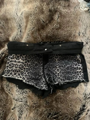 Leopardmönstrade shorts med bälte - Säljer ett par svarta shorts från Bik Bok med leopardmönstrad framsida och råa kanter. Shortsen har ett brett midjeband med knyt och dekorativa nitar. Baksidan är helsvart med två fickor. Perfekta för dig som vill sticka ut!