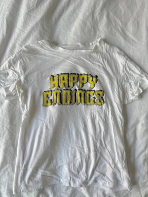 Ganni T-shirt  - Vit t-shirt från Ganni med coolt tryck i gult och svart där det står 'Happy Endings' på bröstet. Klassisk passform och rund halsringning, perfekt till jeans eller kjol.
