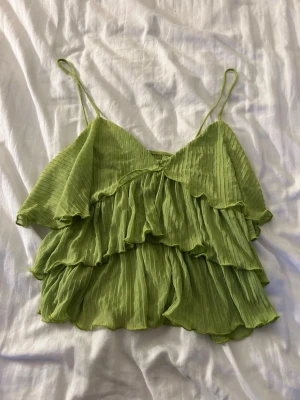 Grön volangtop från Zara - Säljer ett ljusgrön linne från Zara med tunna axelband och flera lager volanger. Toppen är plisserad och har en luftig, flowy känsla som passar perfekt till sommaren. Materialet känns lätt och svalt.
